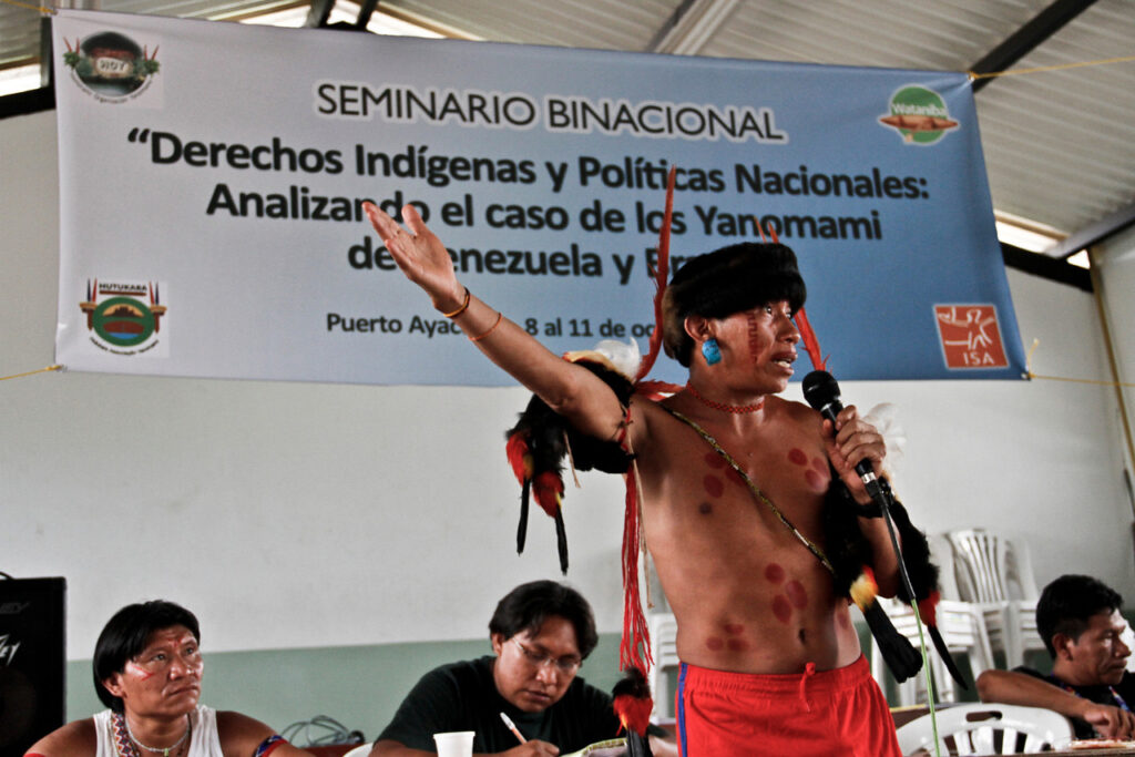 A young Indigenous man stands and speaks, holding a microphone, in front of a large banner announcing a conference on Indigenous rights. His chest is bare, with round red markings in several areas. He has a vertical red design on his visible cheek; a large turquoise-colored earring; a thick black band across his forehead; black, red and white feathers tied to his upper arms; and is wearing red athletic shorts. Three Indigenous men seated at a table are visible behind him.