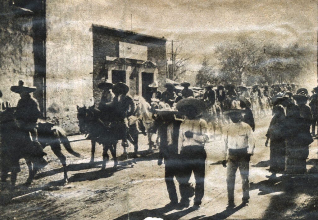 A news clipping in black and white shows a large group of people marching on an outdoor street. Some are on horseback. Almost all wear sombreros. A low building can be seen in the medium background, as well as trees farther back.