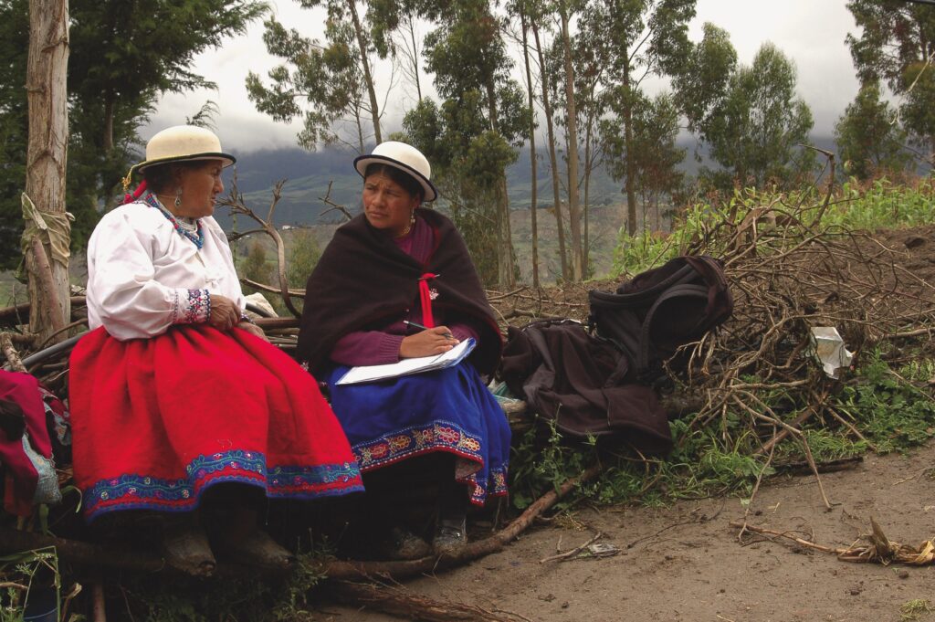 Two Indigenous women sit on fallen tree limbs, in conversation. They both wear long, full skirts, white bowler hats with black rims. The one on the right holds a notepad on her lap and a pen in her hand. She wears a dark brown shawl pinned shut and her skirt is an intense blue with a colorful border. She listens as the other woman speaks. The second woman wears a white long-sleeved blouse with border at the cuff and a bright red skirt with a border of blue and other colors. They are seated in a somewhat cleared area in a forest, along a dirt path. Trees and hillsides are visible in the background, as well as low, heavy clouds.