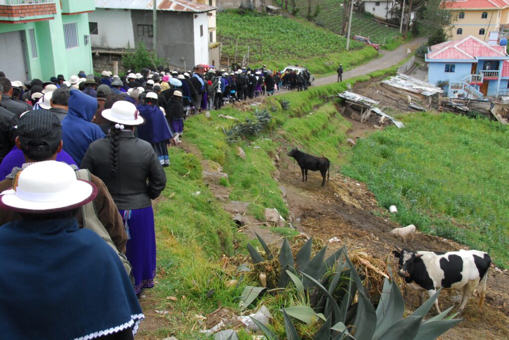 On a path set in bright green hillside, a long procession of people walks in the opposite direction from the camera. Many of them have white bowler hat. Women have long skirts and capes or shawls. T the very front of the procession is a car. There are a few houses visible on either side of the path, painted in pastel colors. Several cows and other animals are also visible, as are cultivated rows of plants.