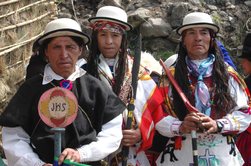 Two men and an adolescent face the camera. All are elaborately costumed in white bowler hats, adorned with several scarves or a cape. A white apron embroidered with various colorful symbols is visible on the man and the adolescent, as are many small, long braids. The adolescent, in the center, holds a sword aloft. The two men rest their hands on the tops of bats or clubs. In the immediate background, a rocky hillside is visible as well as a structure made of thin wooden rods and straw.