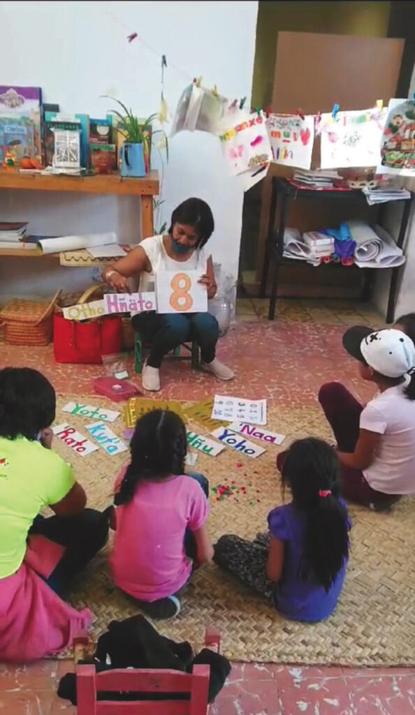 In a children's classroom, a woman with dark hair is seated on a low chair facing a small semi-circle of children. She is holding a square of paper with a large number 8 written on it in orange. She is also holding a card with a word on it in an Indigenous language. On the floor in front of the children are other strips of paper with words on them written in large print and bright colors. Behind the teacher, there is a shelf with books and other materials, baskets, and a string from one wall to another where drawings are hanging from clothespins.