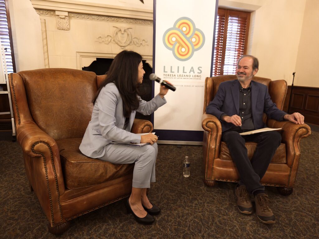 A woman in a pale blue suit with long, straight dark hair sits in a large leather chair and holds a microphone as she faces a man, also seated, smiling in a matching leather chair. He is wearing a dark blue suit jacket, dark blue shirt, and black pants.