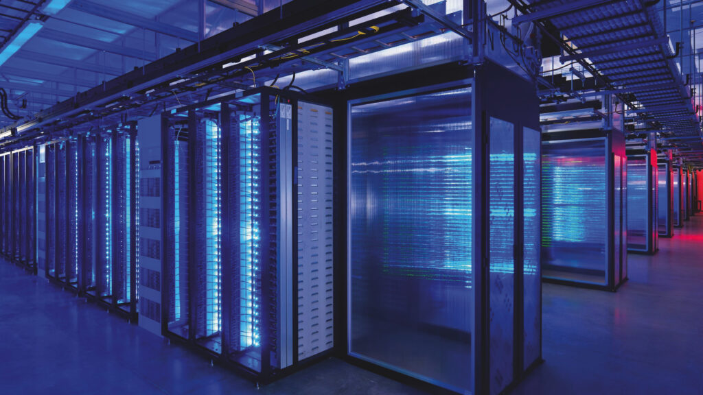 A room that appears almost entirely blue, with rows of tall, rectangular connected glass boxes. Some wiring is visible above them. The room appears to be enormous. Light blue light emanates from many of the compartments and on the right-hand side red light emanates from an unseen source.