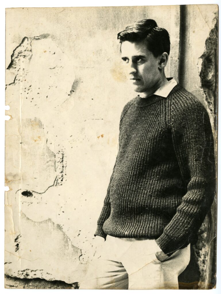 Black-and-white photo of a man leaning against a rough-looking wall, whose plaster is peeling. He has short, dark hair; wears a dark knit sweater with a white shirt collar visible; light-colored pants with his left hand in the pocket. His expression is serious. He is at a 3/4 angle and looks at something out of view.