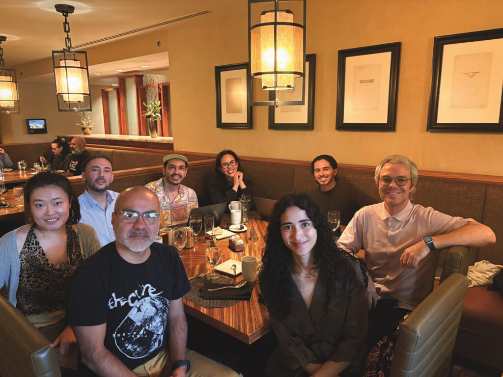 In a restaurant booth with yellowish light, a group of eight people, both men and women, sits around a table, all facing the camera and smiling. Cups, glasses, and silverware can be seen on the table.
