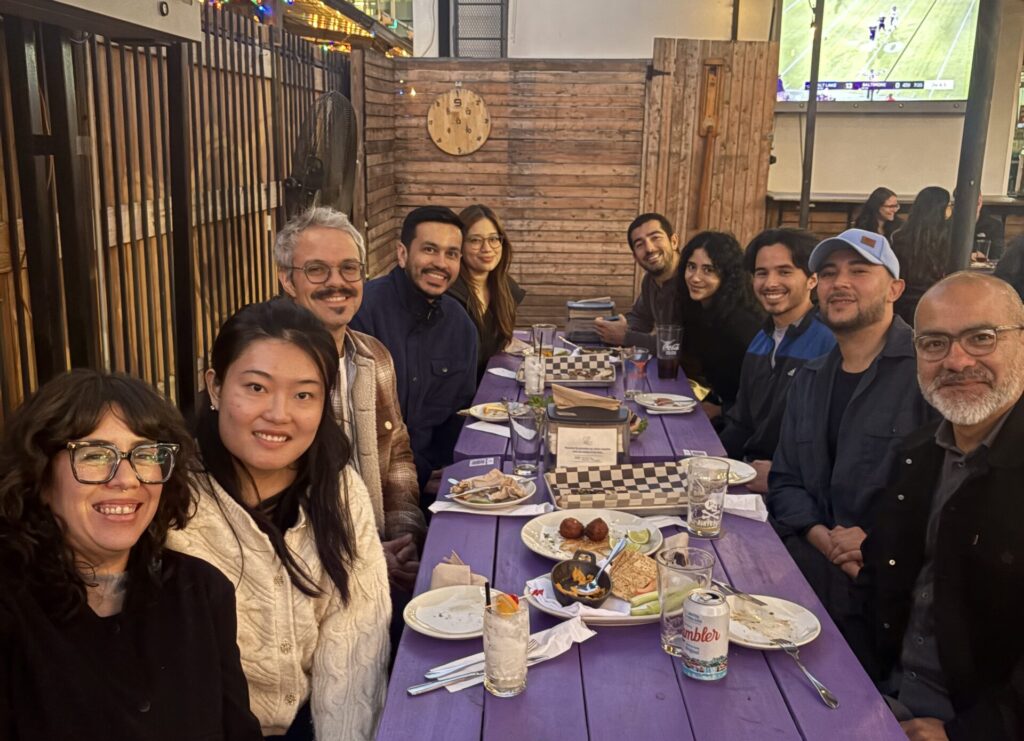 A group of 10 adults, both men and women, sits at two long wooden tables at an outdoor restaurant. The tables are picnic tables painted purple. There are plates and glasses visible. The group smiles at the camera.
