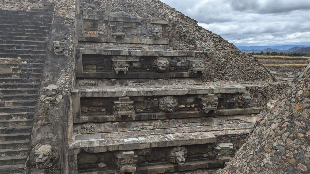 In the foreground is the side of a stone pyramid with smaller steps to the left, and larger tiered platforms to the right. There are animal heads and other carvings jutting out along the vertical edge of the stairs as well as the horizontal platforms. Masses of smaller stones are visible along the sloped sides of the structure. In the distance, a cloudy sky floats above mountain ranges that are blue and dark brown, as well as a small but distant row of dark green trees.