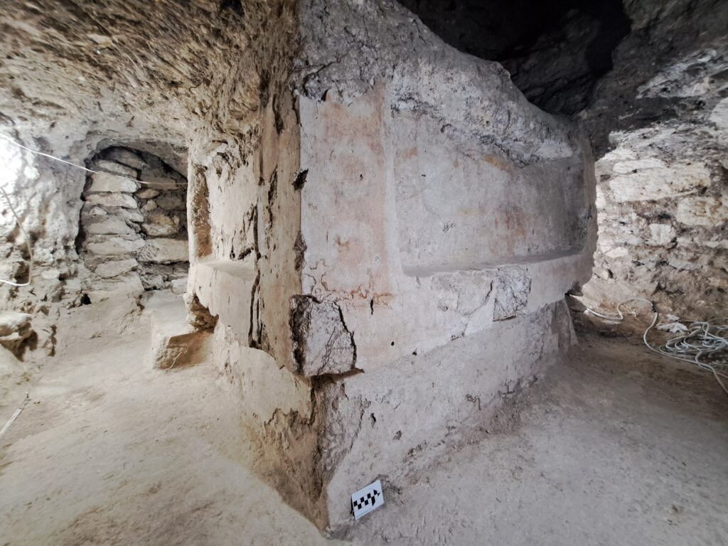 Image shows crumbling stone walls and passageways of whitish color that appear to be the interior of an almost cave-like structure. The large wall in the foreground shows faint evidence of having been covered with stucco and painted with reddish coloring.
