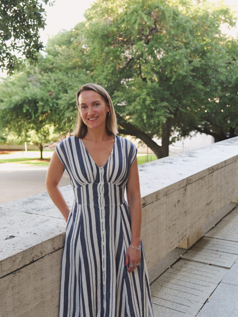 A white woman with shoulder-length straight blondish hair stands in a blue and white striped dress, facing the camera and smiling. She is outdoors, standing along a waist-level limestone wall. In the distance there is a large live oak tree.