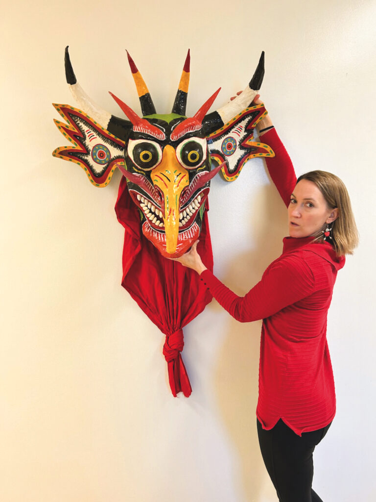 A white woman with blond shoulder-length hair holds a large mask that is mounted on the wall. She is wearing a bright red sweater, red and white earrings, and black pants. The mask is a large devil's face with six horns, a long yellow nose and bulging black and yellow eyes. The mouth is smiling with very prominent rows of teeth. Other colors on the mask are red, white, blue, and a bit of green. A bright red cape that is knotted at the bottom hangs from the mask.
