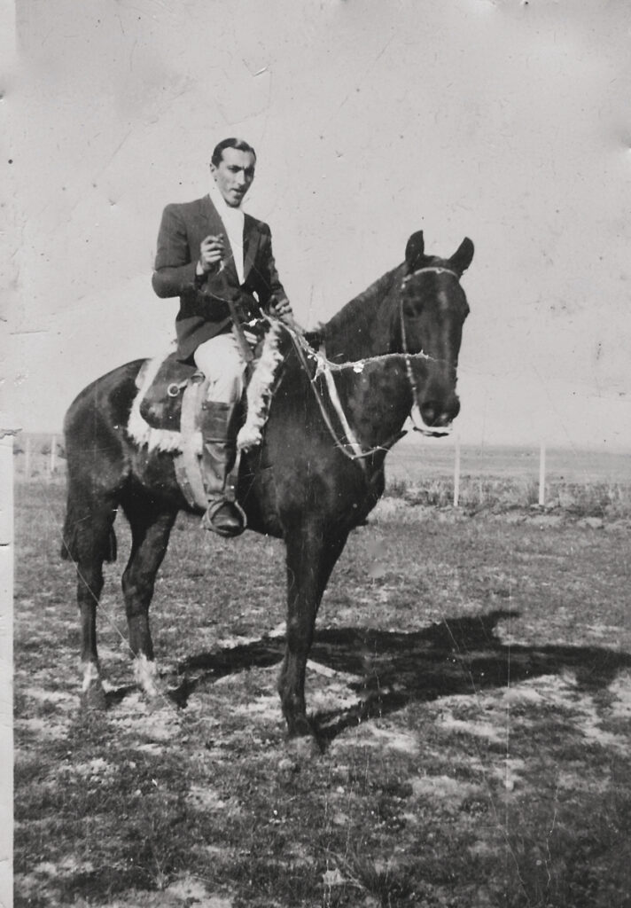 In an aged black-and-white photograph, a man sits on a dark horse in a wide open field. He his wearing a dark jacket and boots, pale shirt and pants, and holds a small riding whip in one hand. His hair is dark and slicked back from his face.