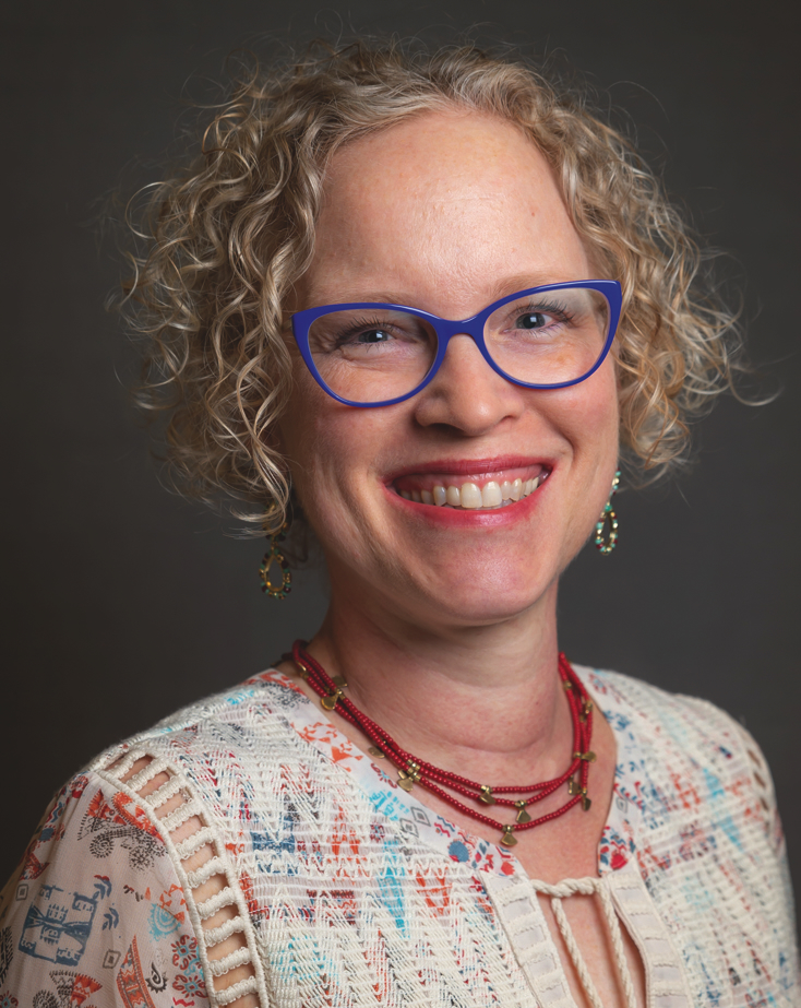 A white woman with blond curly hair and blue eyes smiles broadly. She has bright blue-rimmed glasses, wears a red beaded necklace with bronze figures on it, and shiny gold and turquoise earrings.