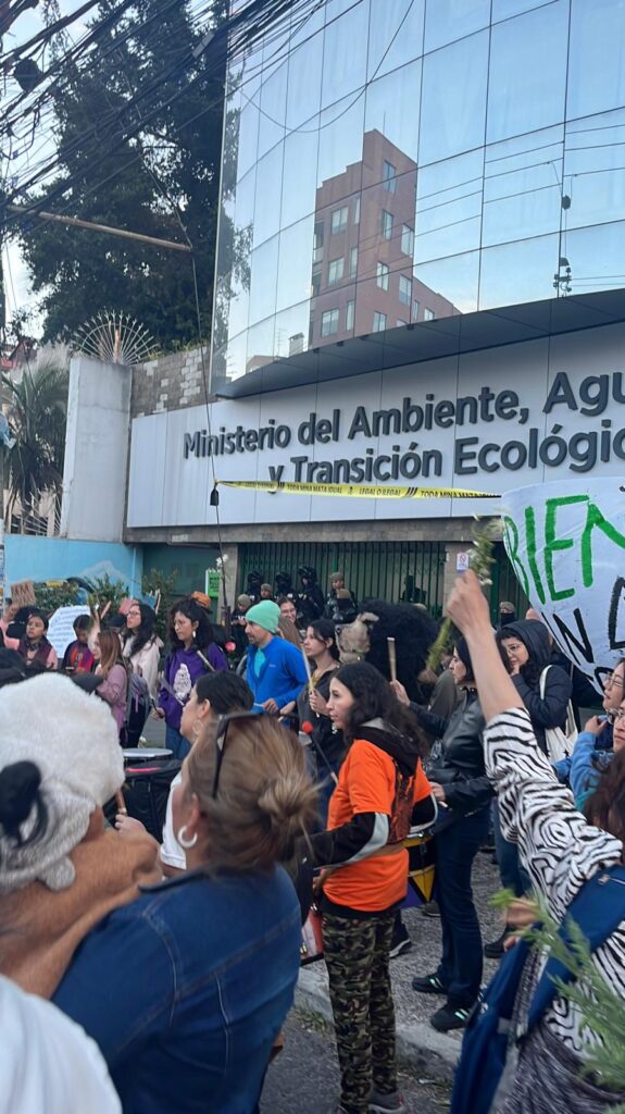 A group of people stand outside the office of Ecuador's Ministry of the Environment, Water, and Ecological Transition. One person holds a protest sign, others hold drumsticks and are beating drums.