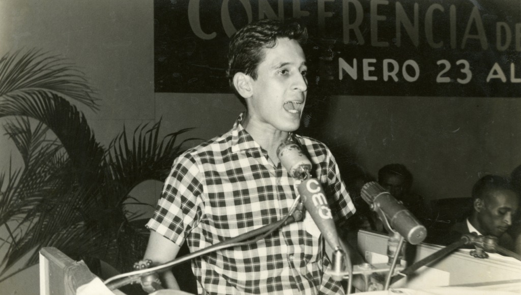 Black and white photo. A white man with short dark hair is at a podium, his mouth wide open as he speaks into a microphone. He is wearing a short-sleeved plaid shirt. In the background, the heads of a two seated people can be seen. Behind, on the wall, is a large banner in Spanish announcing a conference.