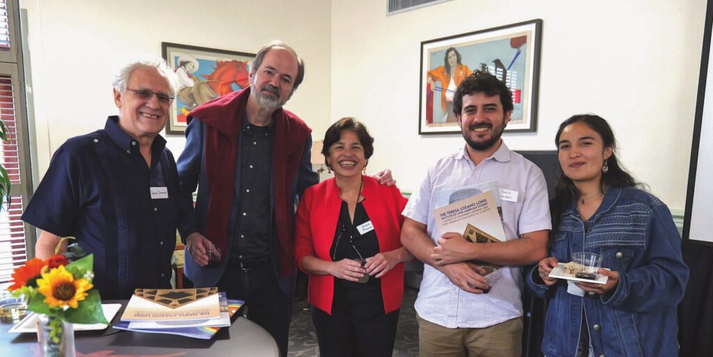 Five adults pose for a photo. Mexican author and public intellectual Juan Villoro stands tall in a dark blue shirt and jacket with a burgundy sweater around his neck. His arm is on the shoulder of LLILAS Director Adela Pineda, smiling broadly in a bright red jacket and black shirt and pants.