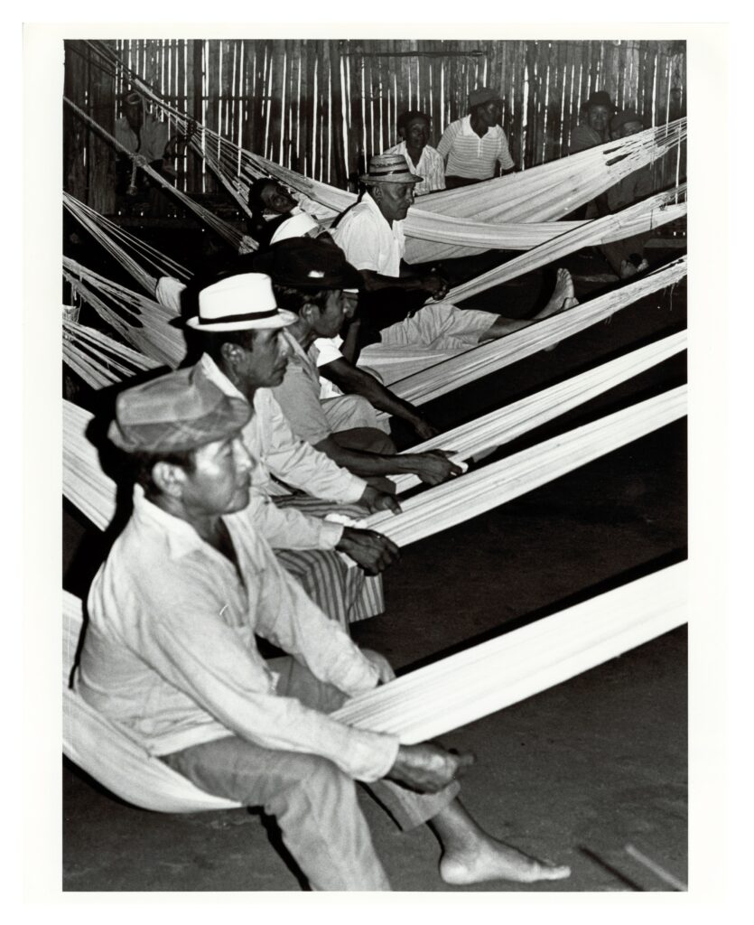 Black and white photograph of a series of seven white hammocks. Men sit astride them, sitting in profile. They are inside a structure with walls of vertical pieces of wood or bamboo.