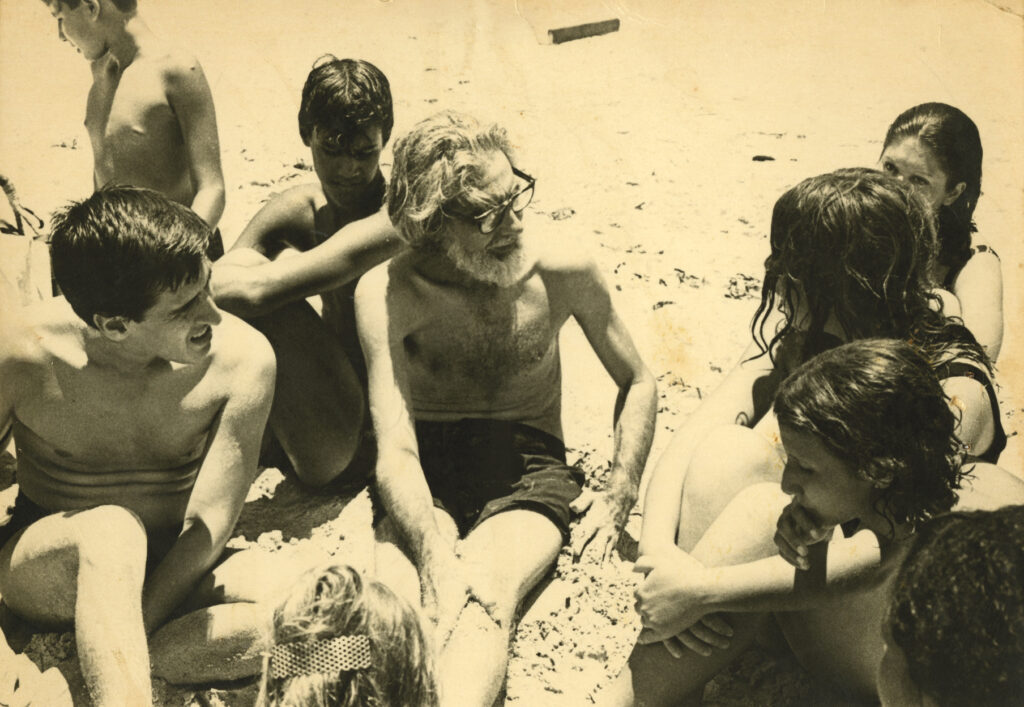 A yellowing black-and-white photo shows a group of people, including children, seated on the sand at the beach. They are all in bathing suits, some with wet hair. In the center of the photo is the late Nicaraguan poet and political activist Ernesto Cardenal, who has a full head of white hair, and white beard and mustache, and dark-rimmed glasses. Next to Cardenal, at the far left, is Roque Dalton, with short dark hair, looking toward a woman who is speaking on the right. He is squinting in the sun.