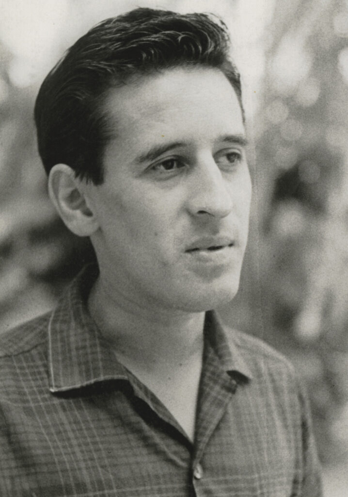 A vertical black-and-white portrait of a young man with pale skin, short, dark hair, a prominent nose. His lips are somewhat parted. His head is three-quarters toward the camera. He is wearing a plaid shirt, and the image extends just below the shoulders.