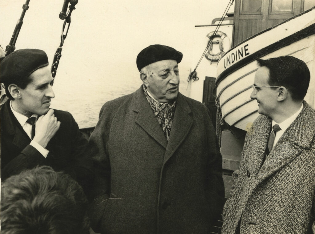 Horizontal black-and-white photo. Three men stand on the deck of a ship. Water can faintly be seen in the background, and in the closer background, the side of a smaller ship with the name UNDINE written in capital letters. Roque Dalton is at left in a dark coat, white shirt, dark tie, and dark beret. He is almost completely in profile, with a had to his chin, and appears to be listening to a conversation. In the middle is an older man, Miguel Ángel Asturias, in a black beret, gray overcoat, and a silken printed scarf tucked in at the neck. He is speaking and facing the man on the far right, who is young, smiling, with short, dark hair, a tweed overcoat, white shirt and striped tie, wire-rimmed glasses.