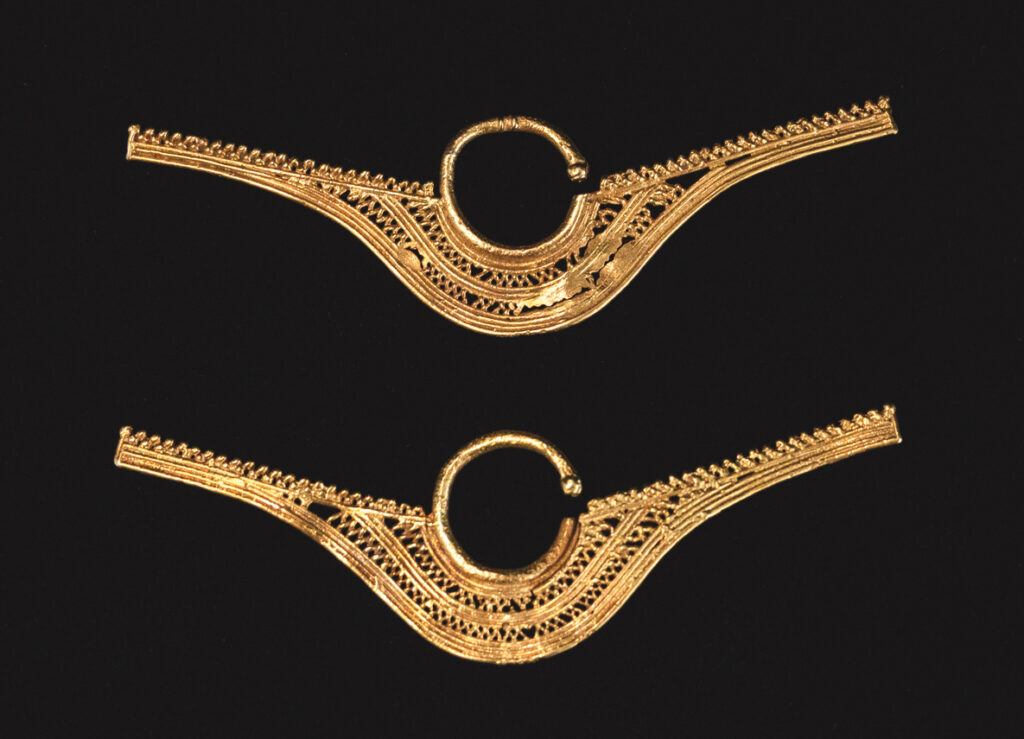 A pair of elaborate bow-shaped gold earrings lies one above the other on a black background. The center is a round circle of gold with an opening for the earlobe. The bow shape is created from long sections of gold wire that are curved as well as smaller ornate sections that resemble filigree.
