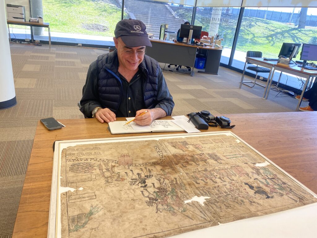A man sits drawing at a table in a large room. In front of him is an old, yellowed document that is several square feet in size, on degraded paper with drawings on it showing many human figures and other shapes. Some colors are visible in the drawing. The man is smiling as he draws similar figures in a small sketchbook in pencil. He is wearing a dark-blue cap, dark blue shirt, and a dark blue puffy vest. In the background, large windows look out on green grass and sunlight. There are tables and desks with computers in the background, and a barely visible person sits at a desk.