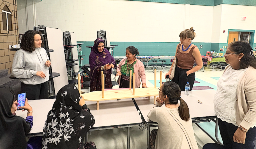 In a large room, a group of women are gathered around two tables. At center, a woman in a headscarf smiles as she stands at a wooden loom that is on top of the table in front of her. She holds strands of thread in her hands. Next to her, a woman smiles and gestures, watching the first woman. She is wearing a richly woven green shirt and a large necklace. Other women are standing or seated, all paying attention to the two at the loom.