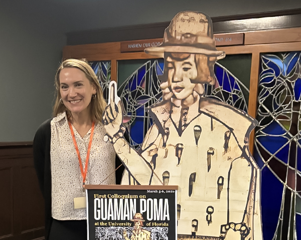 A smiling blond woman stands next to a large cardboard cutout of a man in Spanish colonial garb.