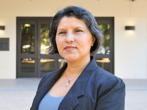 Headshot of Andrea Melendez standing outside of the Walker Mechanical Engineering building on UT campus. Andrea wears a grey blazer, blue blouse, and silver pendant necklace.