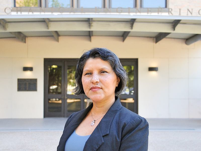 Headshot of Andrea Melendez standing outside of the Walker Mechanical Engineering building on UT campus. Andrea wears a grey blazer, blue blouse, and silver pendant necklace.