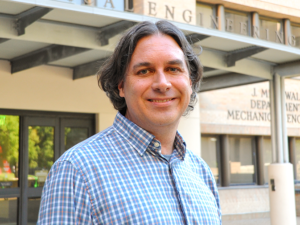 A headshot of John Cassibry standing outside of the Walker Department of Mechanical Engineering building on UT campus. John wears a blue, white, and yellow gingham checked button down shirt.