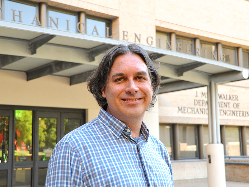 A headshot of John Cassibry standing outside of the Walker Department of Mechanical Engineering building on UT campus. John wears a blue, white, and yellow gingham checked button down shirt.