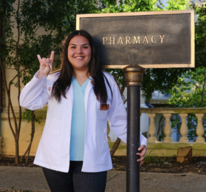 Joann Hi giving hook 'em horns hand sign and standing next to pharmacy building sign.