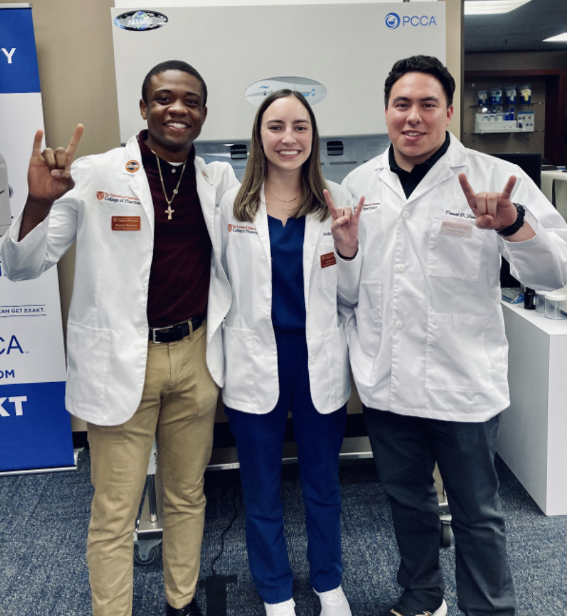 Three members wearing pharmacy white coats and giving hook 'em horns hand sign.