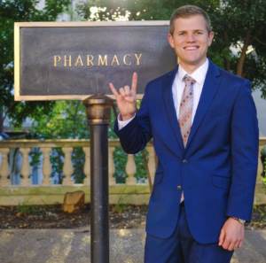 Weston Esplin wearing a blue suit and giving hook 'em horns hand sign in front of pharmacy building sign.
