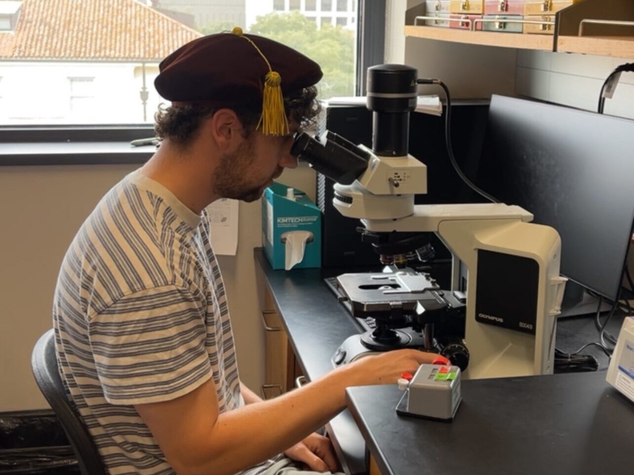 Ryan using a microscope and wearing a graduation hat.