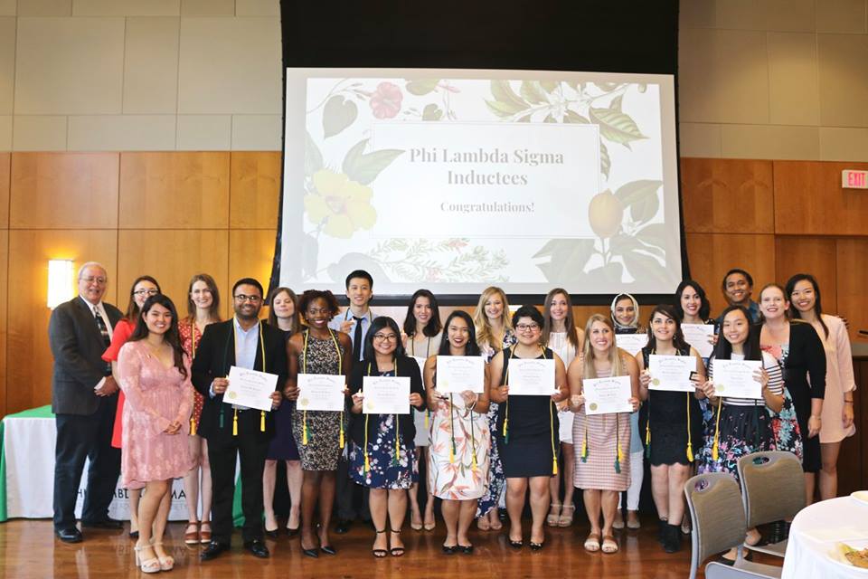 Group of students with faculty advisors standing in front of projection screen; many students are holding up certificates and wearing honor cords.