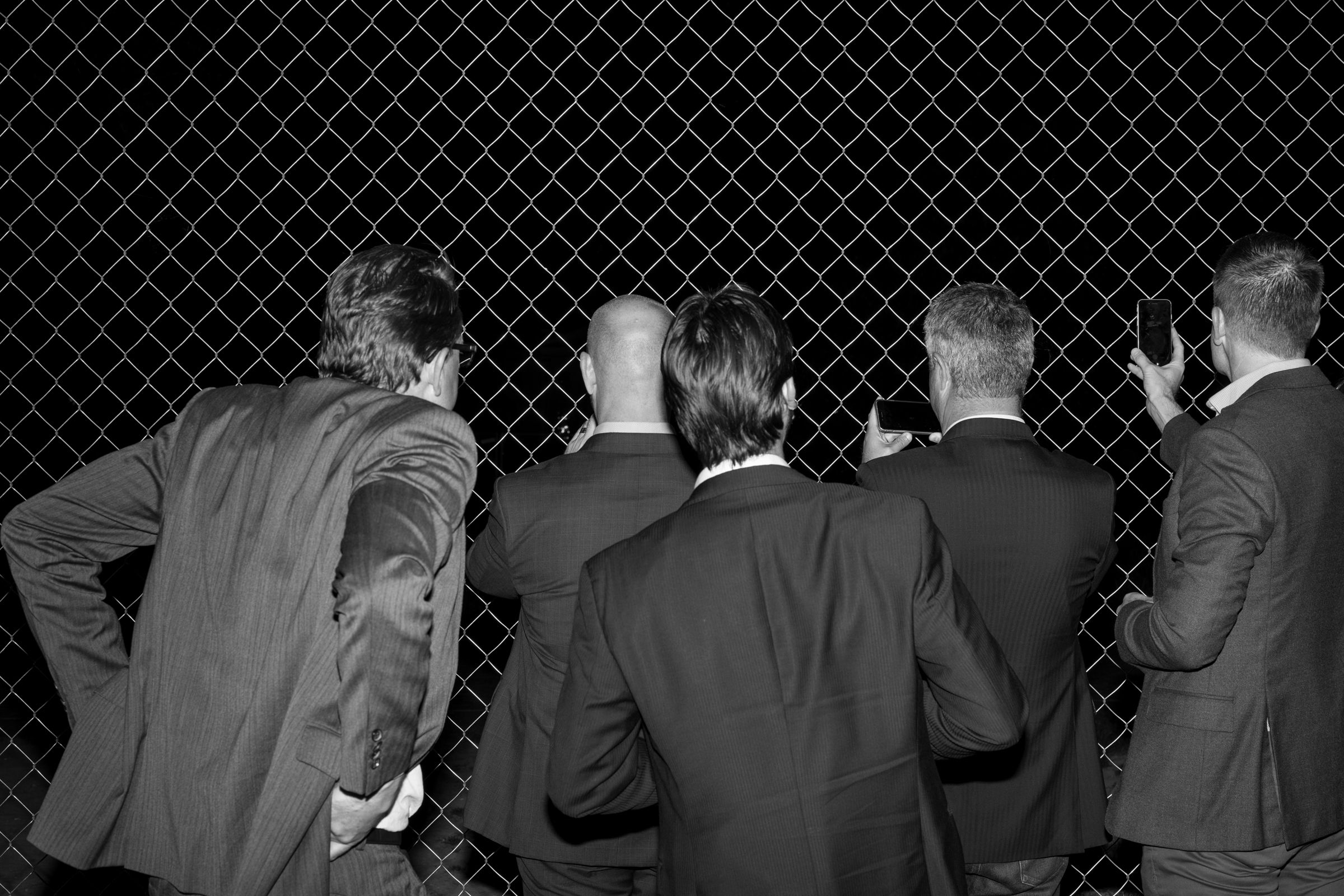 five mens in suits taking pictures with cellphones through chainlink fence