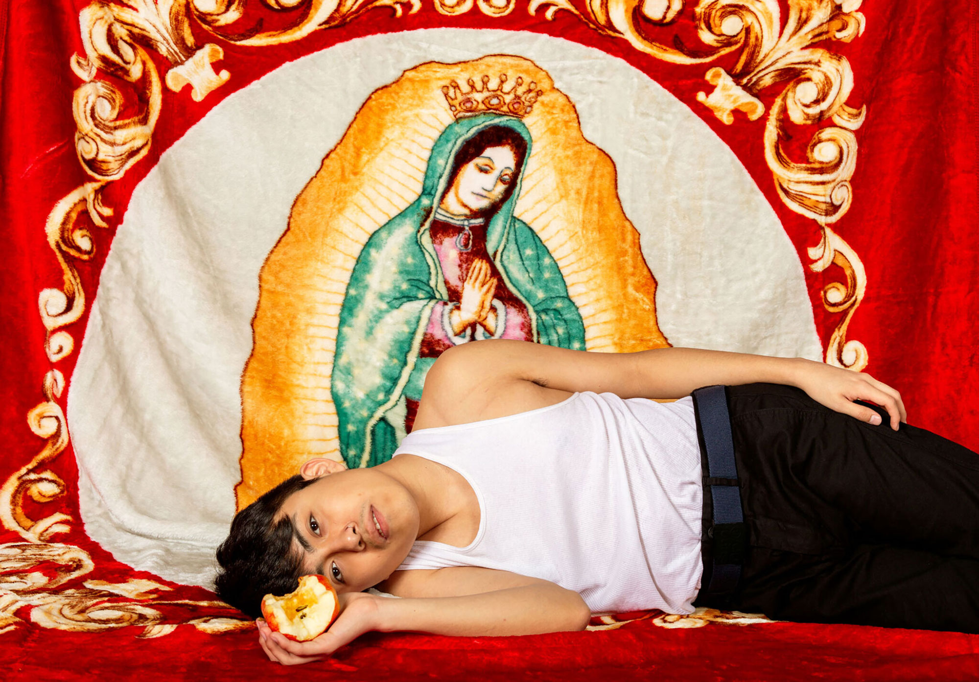 a child holding a partially eaten apple and laying on side in front of textile displaying image of Virgin Mary