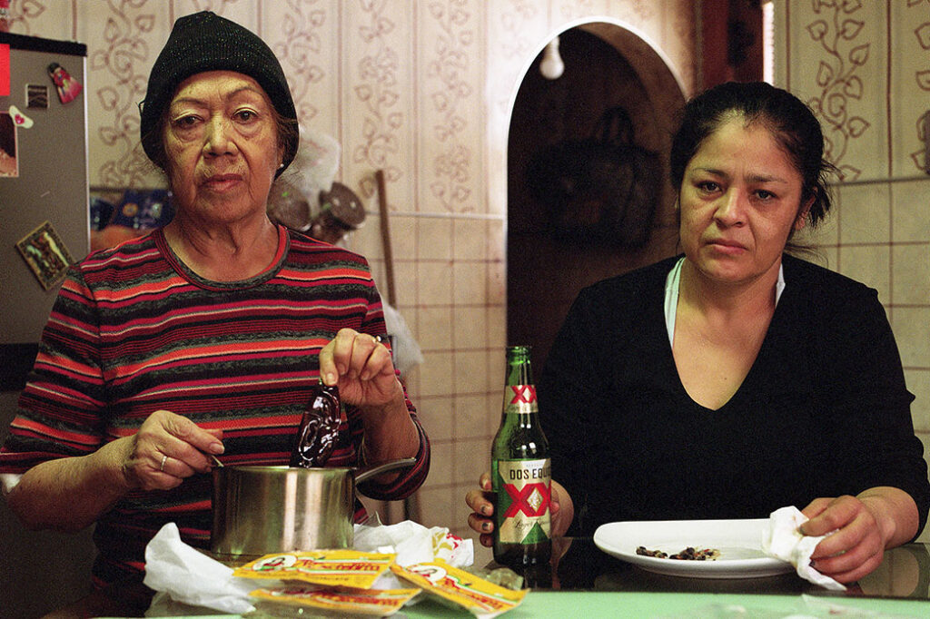 two adults staring directly at camera, in room that might be a kitchen, with one holiding a spoon in a pot, and the other holding a beer bottle next to an empty plate