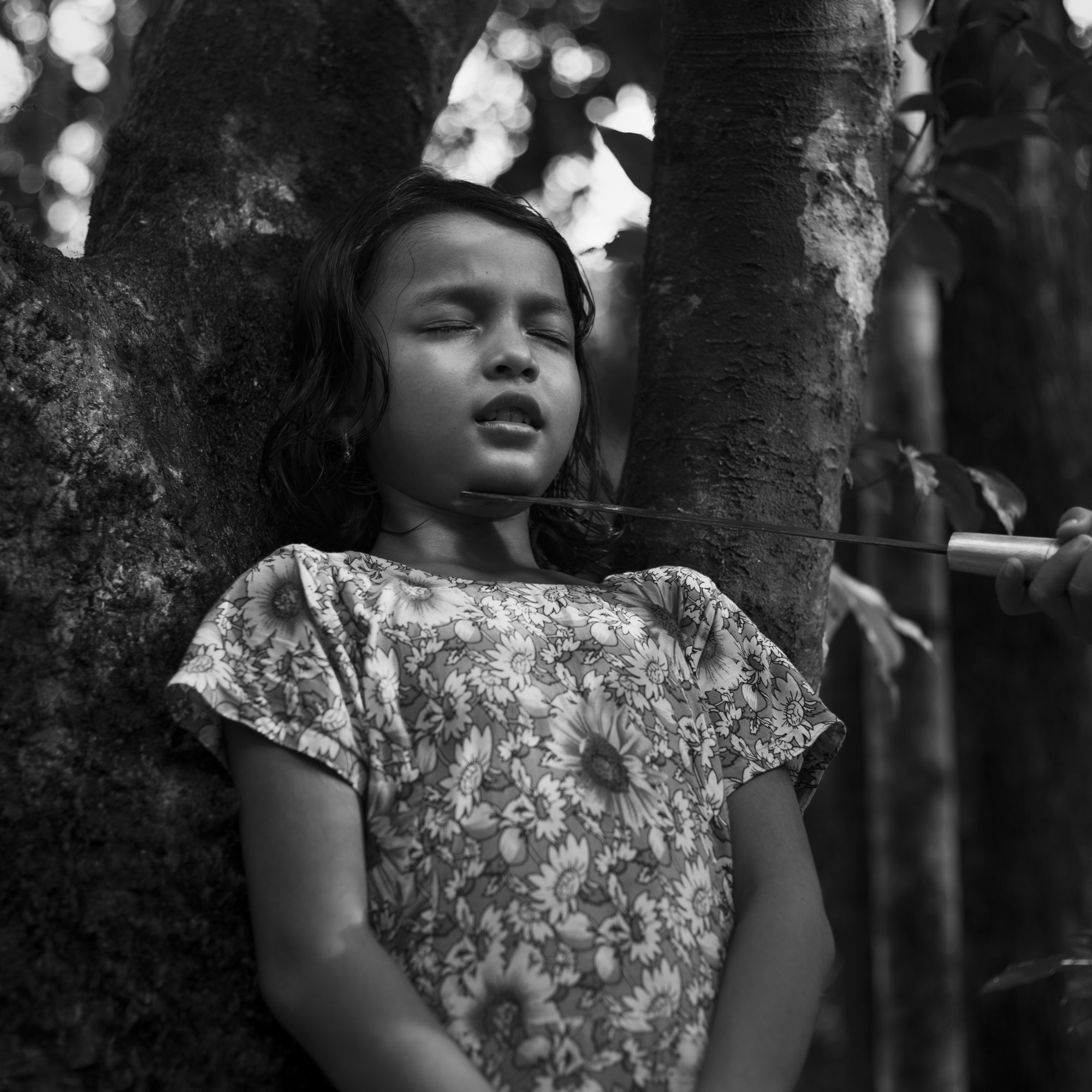 child leaning against tree while hand at edge of frame holds rod that is touching their chin