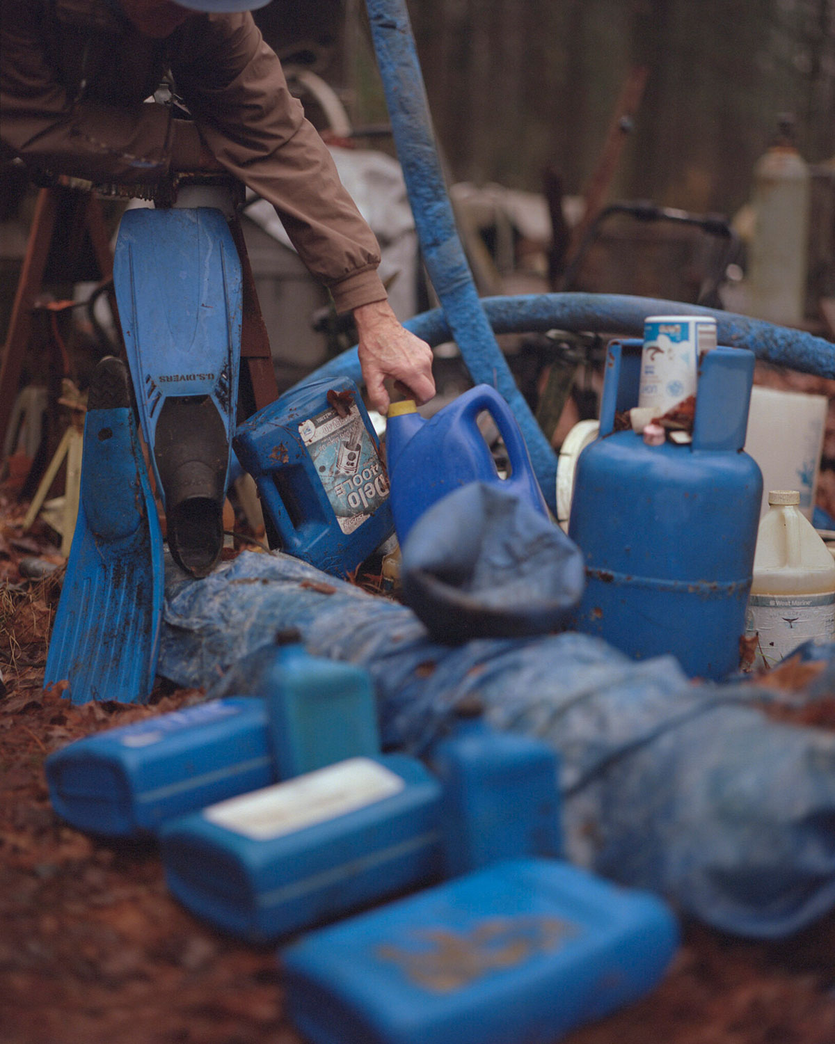 human arm reaching down and holding the lid of a blue jug, which is surrounded by other blue items in what appears to be a junkyard