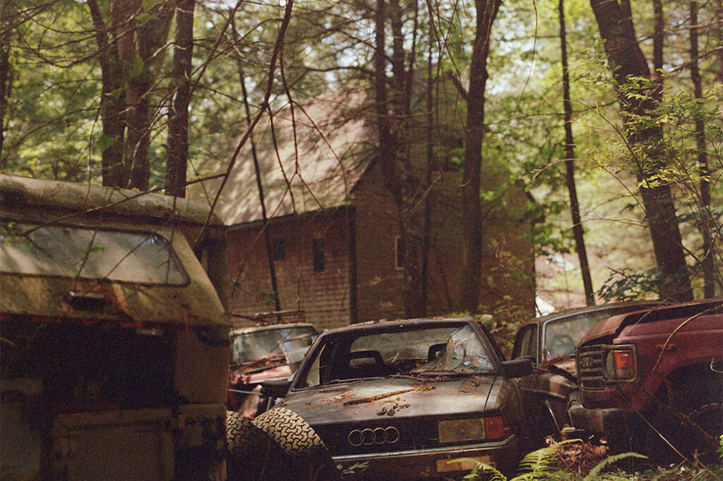 detail of dilapidated Audi car among other dilapidated vehicles, all within a wooded area surrounding a house behind them