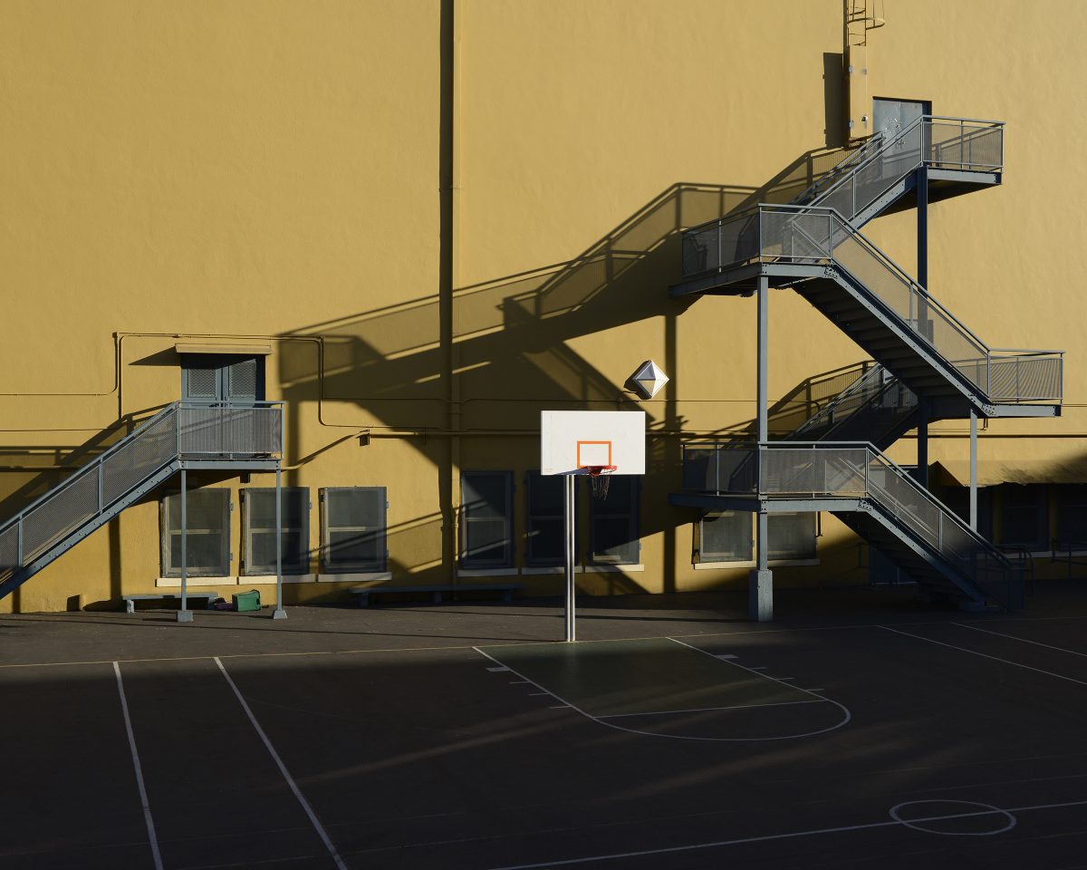 basketball goal on court in front of yellow building with grey fire escape stairways extending beyond doors