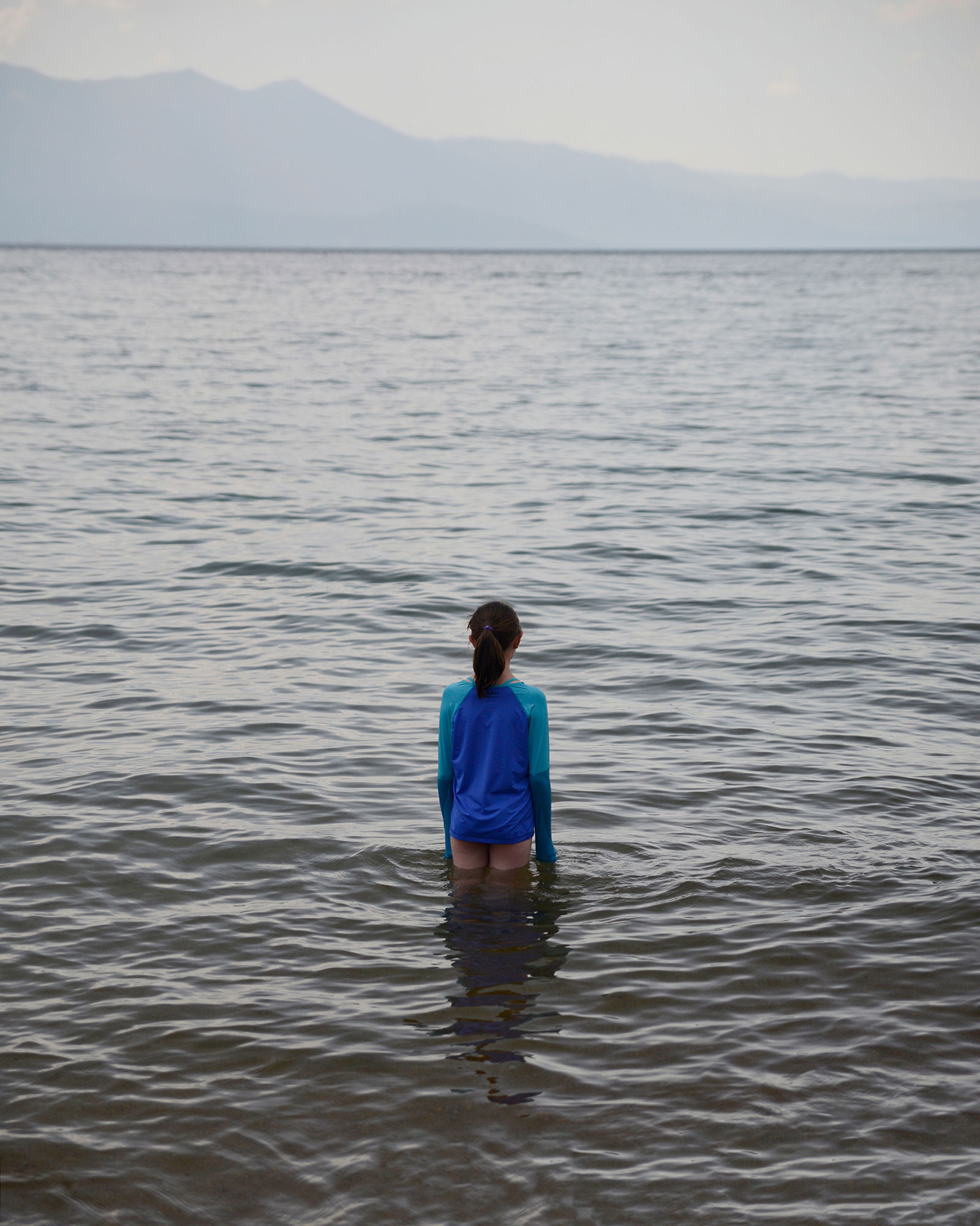 human figure standing in waist-deep water of ocean and wearing a blue and teal long sleeve shirt