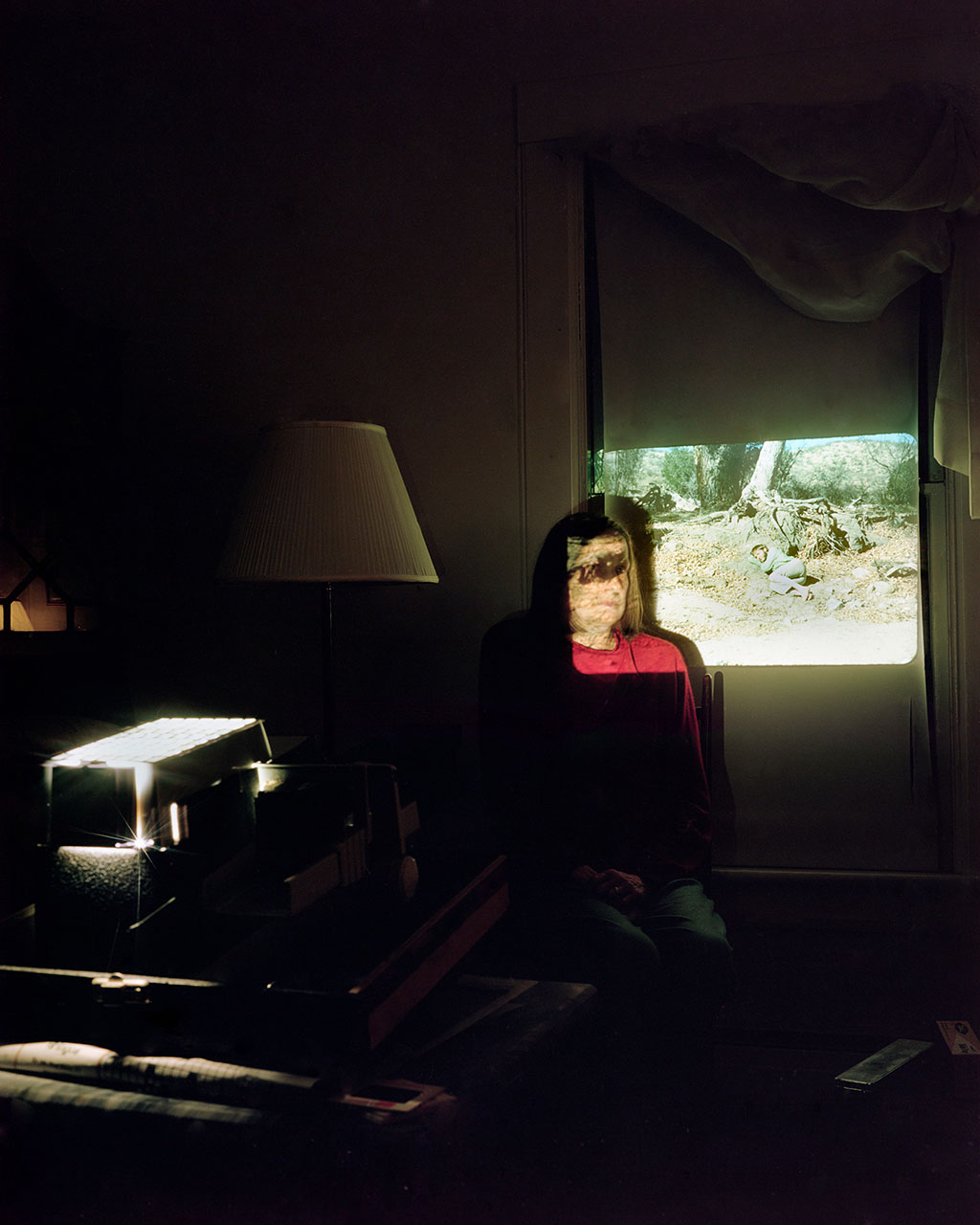 person sitting against wall of dark room with film projector casting light against their face and wall