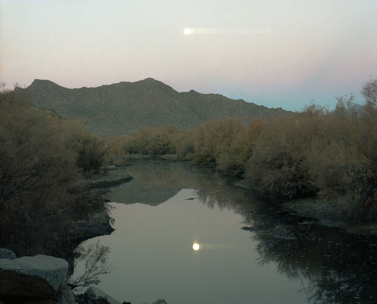 moon at early dusk reflected in the surface of a river with scrub brush on both banks