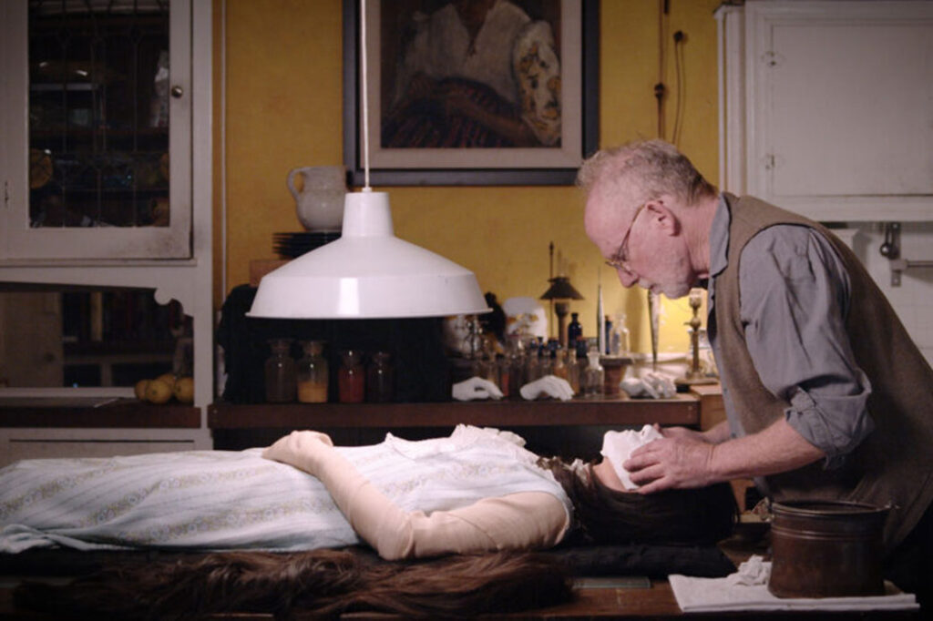 person standing over the head of a human figure laying on a table under a light fixture, with a plaster mask over the figure's face and several plaster casts of hands on as table in the background