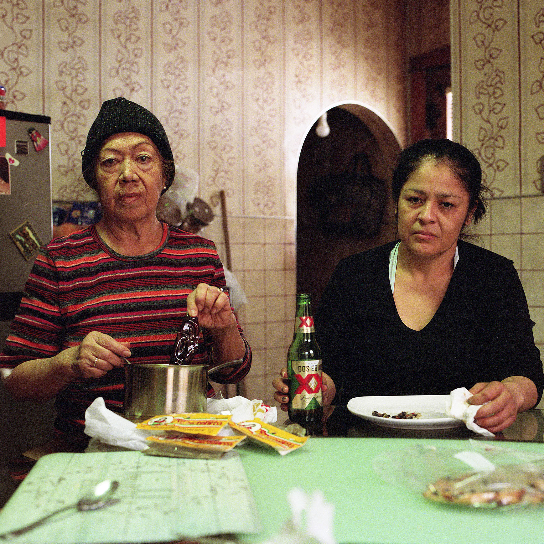 two adults staring directly at camera, in room that might be a kitchen, with one holiding a spoon in a pot, and the other holding a beer bottle next to an empty plate