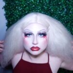 Person with dramatic white & red makeup facing camera with hands and white wig framing face.